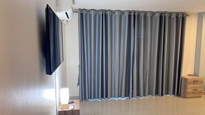 A room with a flat-screen TV mounted on the wall, a curtain, a wooden nightstand with a lamp, and a tiled floor. - The Freelancer Hotel (Pattaya)