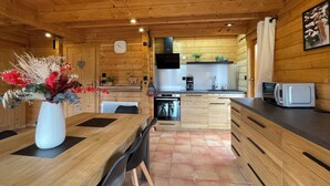 A wooden cabin interior with a dining table, chairs, a kitchen with modern appliances, and a clock on the wall.