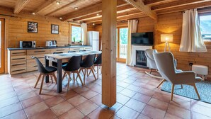 A wooden cabin interior with a dining area, kitchen, and seating by a fireplace.