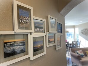 A wall with multiple framed photographs of coastal scenes, including a pier and sunset views. - Sea Breeze with Ocean View Balconies | 3BD/2.5BA (Pacifica)
