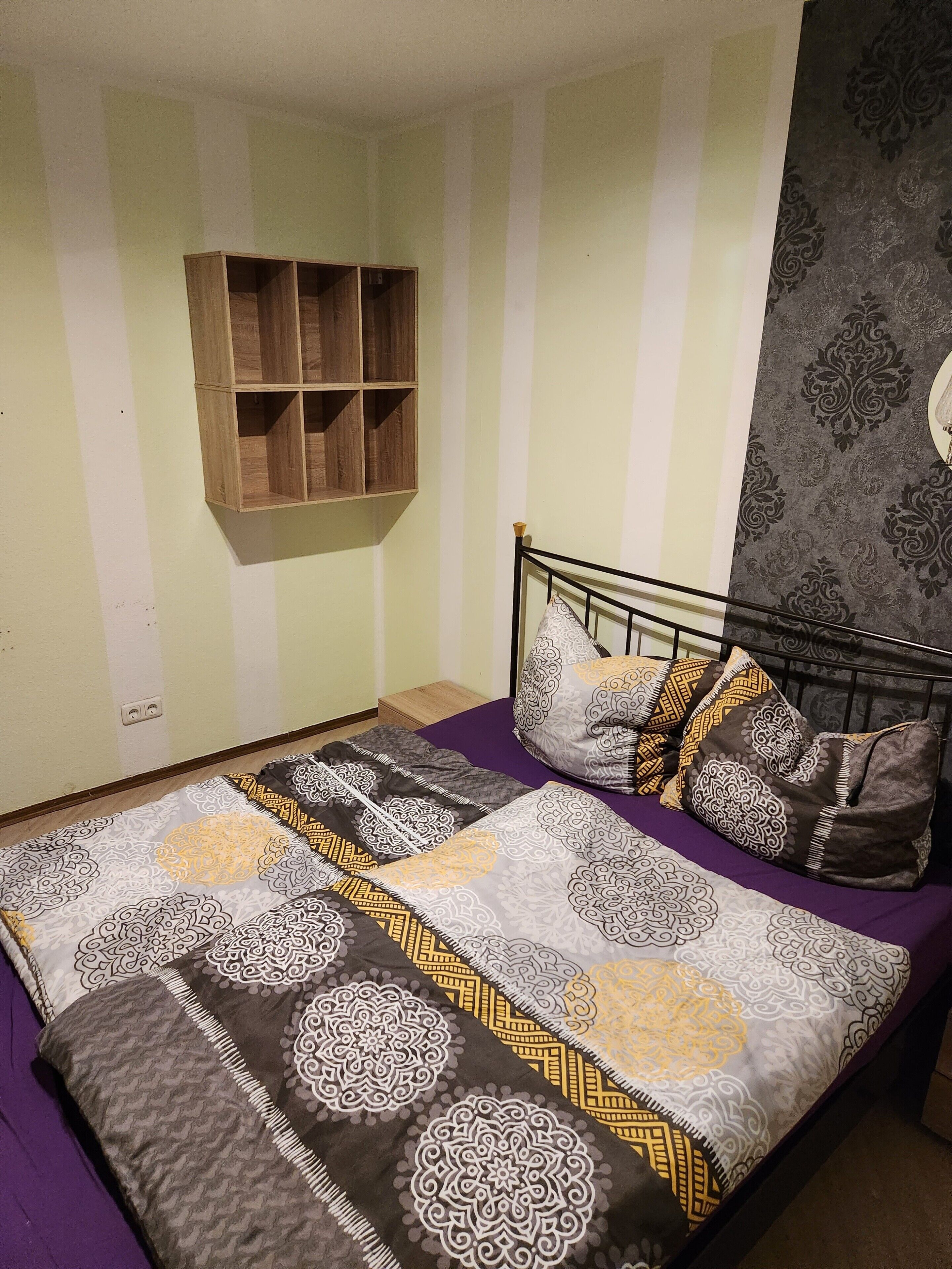A bedroom with a bed, pillows, a wall-mounted shelf, and a patterned wall.