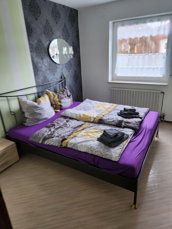 A bed with a purple bedspread, a mirror on the wall, and a window with a view of buildings.