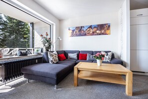 Ein modernes Wohnzimmer mit einem Ecksofa, einem Holztisch, einer Heizung und einem großen Fenster mit Blick auf Bäume.