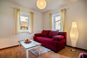 Un canapé rouge, une petite table avec des fruits et une lampe dans une pièce dotée de fenêtres et offrant une vue sur les bâtiments à l’extérieur.