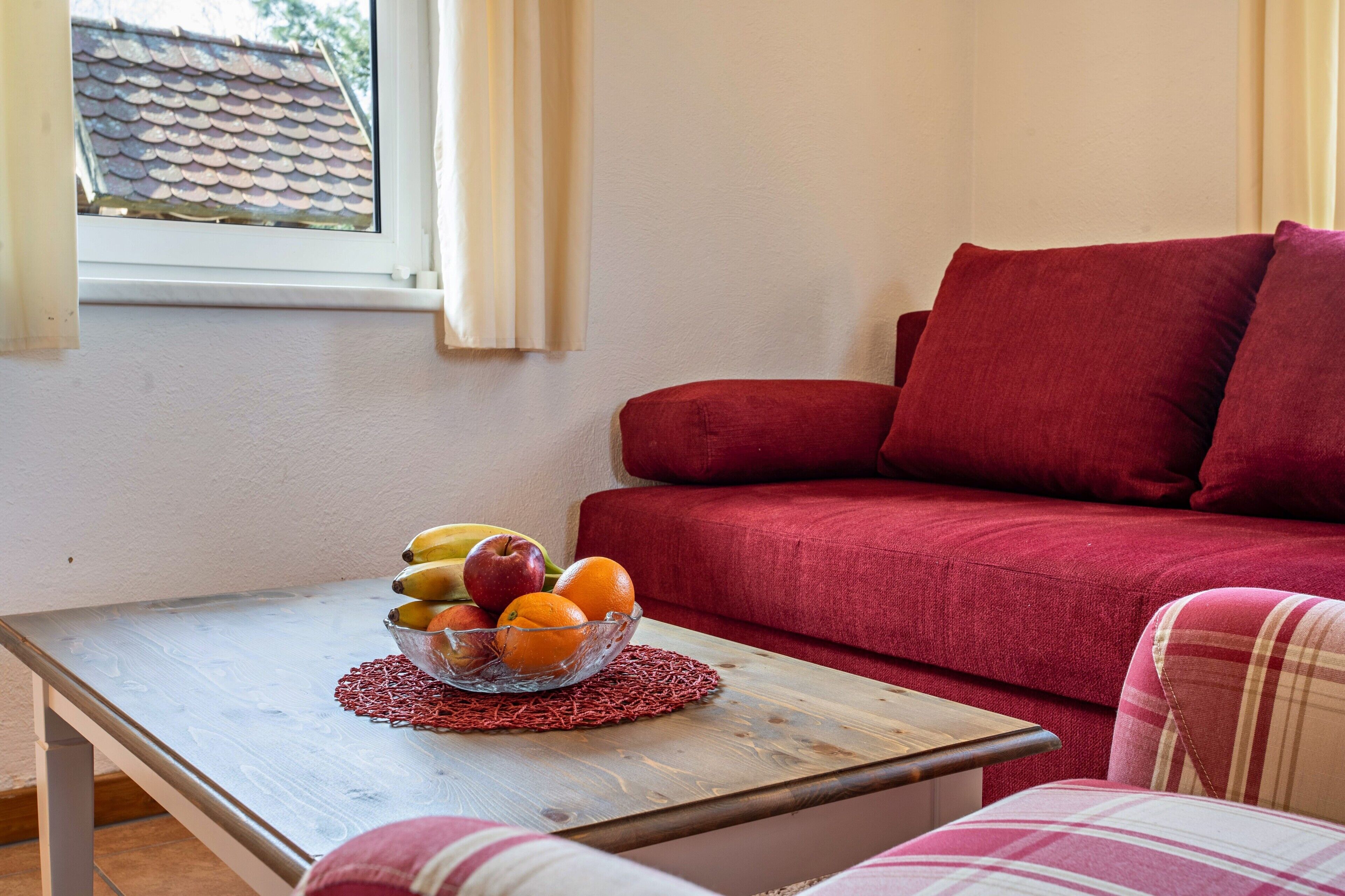 Ein Zimmer mit einem Holztisch, einer Obstschale, einem roten Sofa und einem Fenster mit Vorhängen.