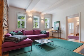 Een woonkamer met een paarse bank, een salontafel en een groen vloerkleed. Er zijn ramen met gordijnen, een schilderij aan de muur en een trap op de achtergrond.
