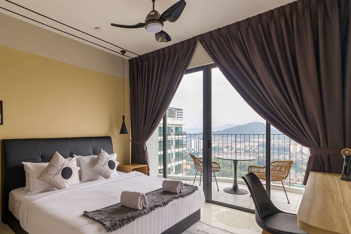Ein Hotelzimmer mit einem großen Bett, einem Deckenventilator, Blick auf die Stadt vom Balkon und einem Essbereich mit Stühlen.
