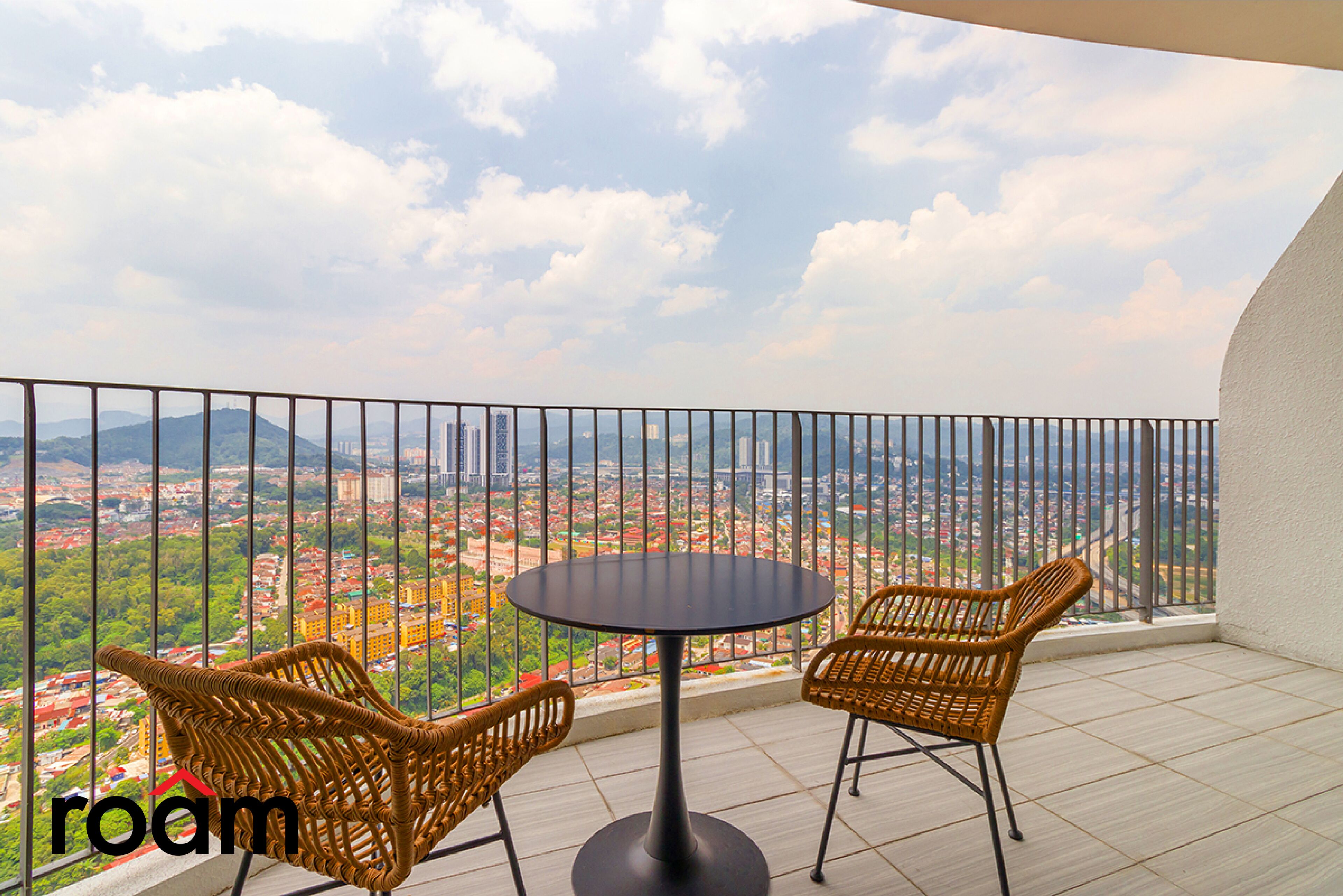 A balcony with a round table and two wicker chairs, offering a view of a cityscape.