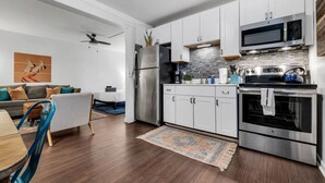 A modern kitchen with stainless steel appliances, white cabinetry, and a dining area with a blue chair and a small table. - Near 12th South | Pool | 3 mi. to Broadway (Nashville)