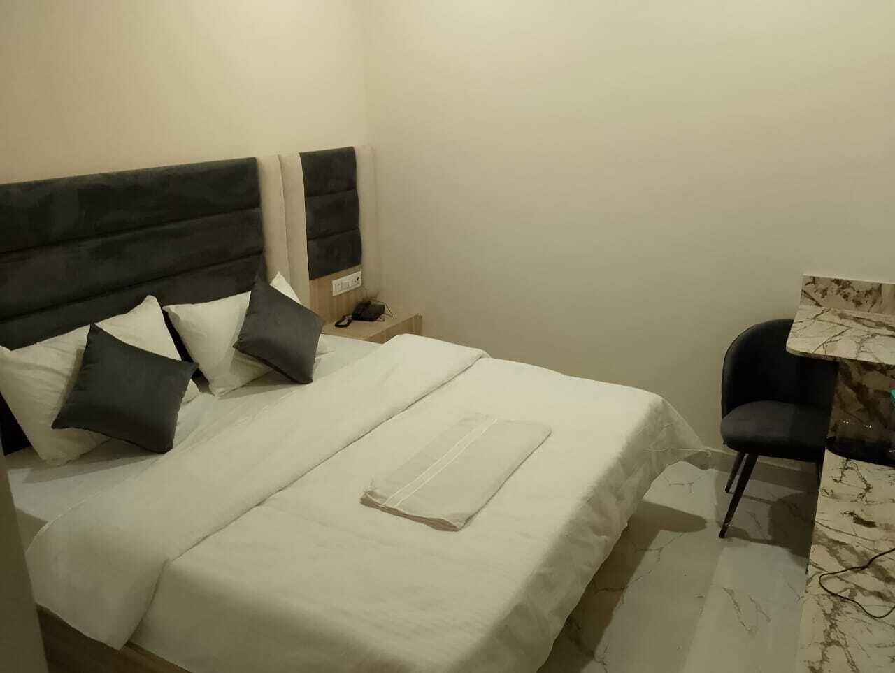 A neatly made bed with white and dark pillows, a bedside table with a phone, and a chair next to a marble-patterned table.
