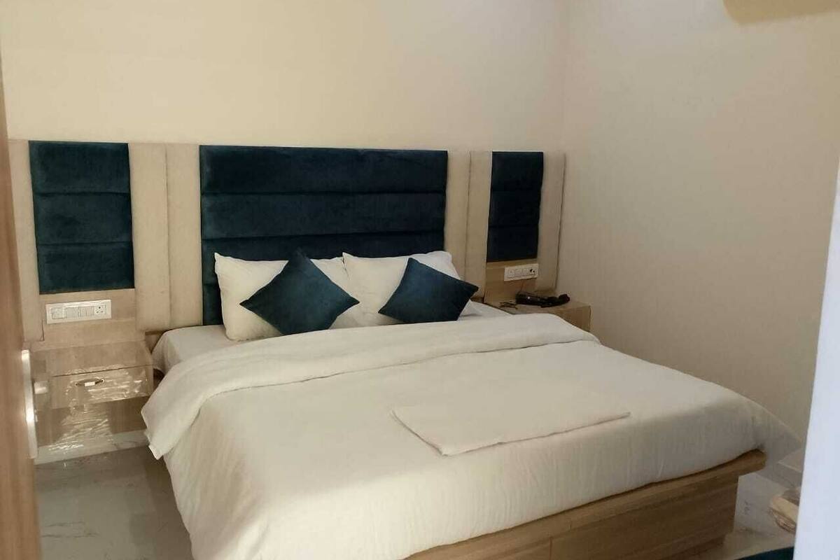 Una cama bien hecha con edredón blanco y dos almohadas oscuras, un cabecero de madera y una mesita de noche con una lámpara.