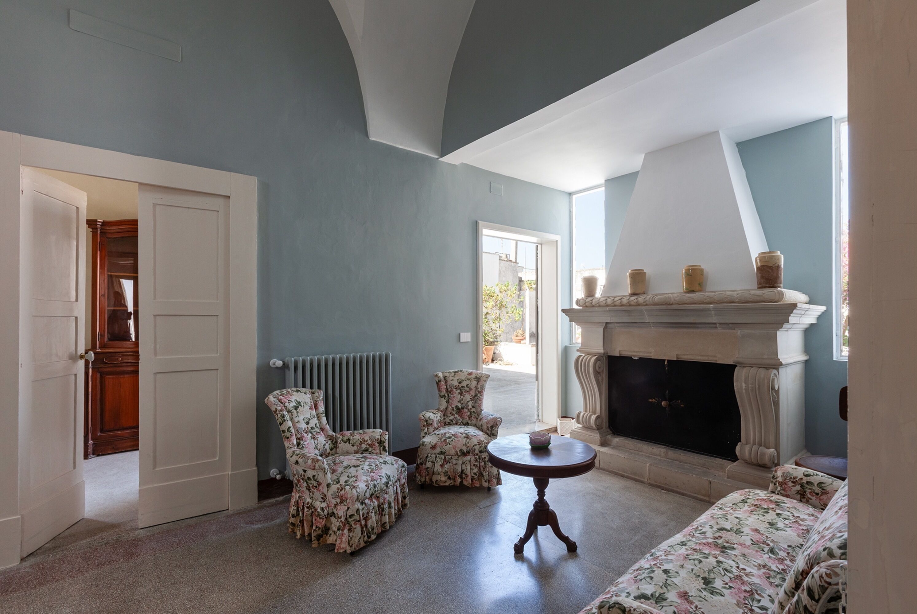 A room with a fireplace, floral armchairs, a round table, and a view of the outside.