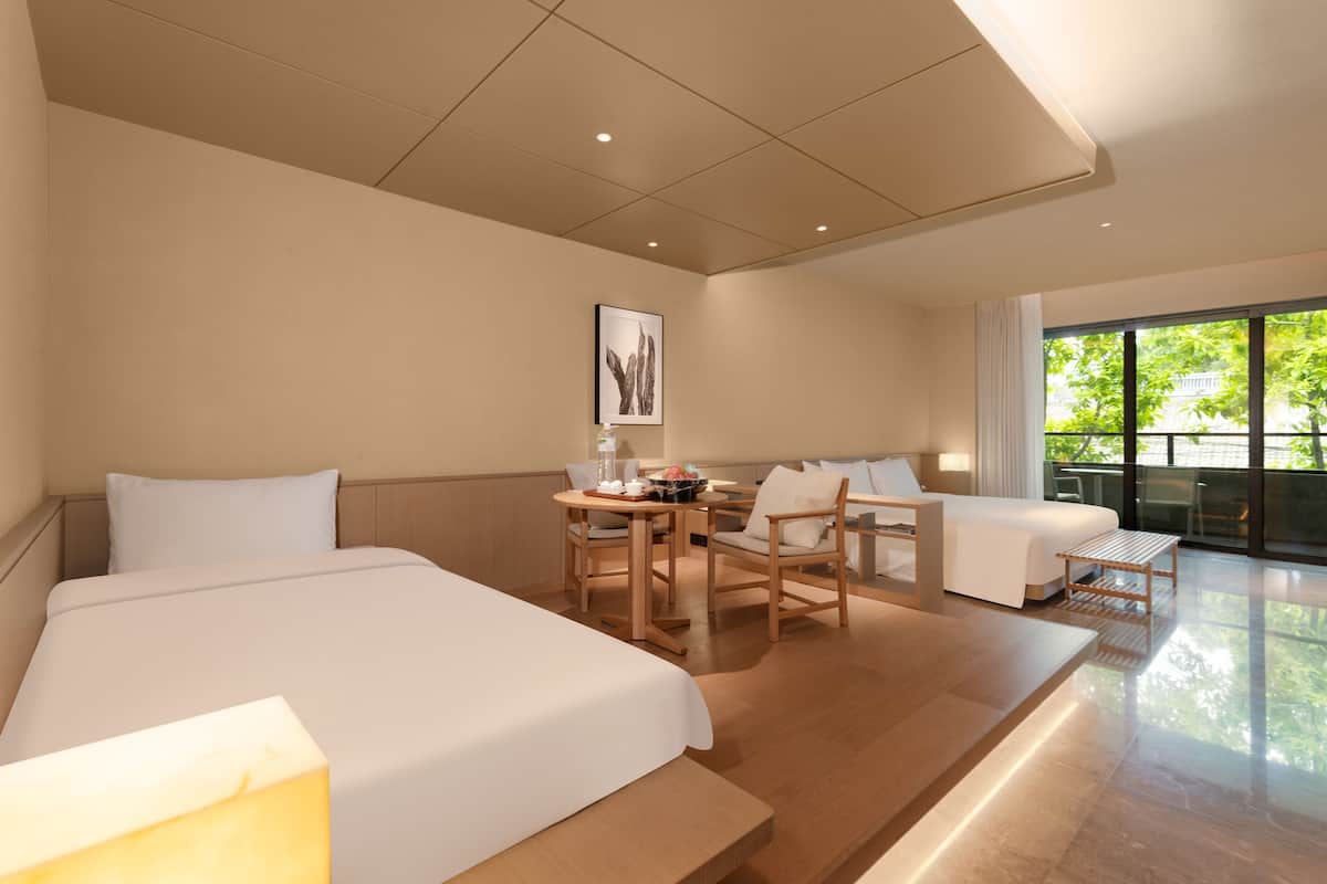 Une chambre d’hôtel avec deux lits, une table à manger, des chaises, un petit canapé et une grande fenêtre donnant sur un paysage verdoyant.