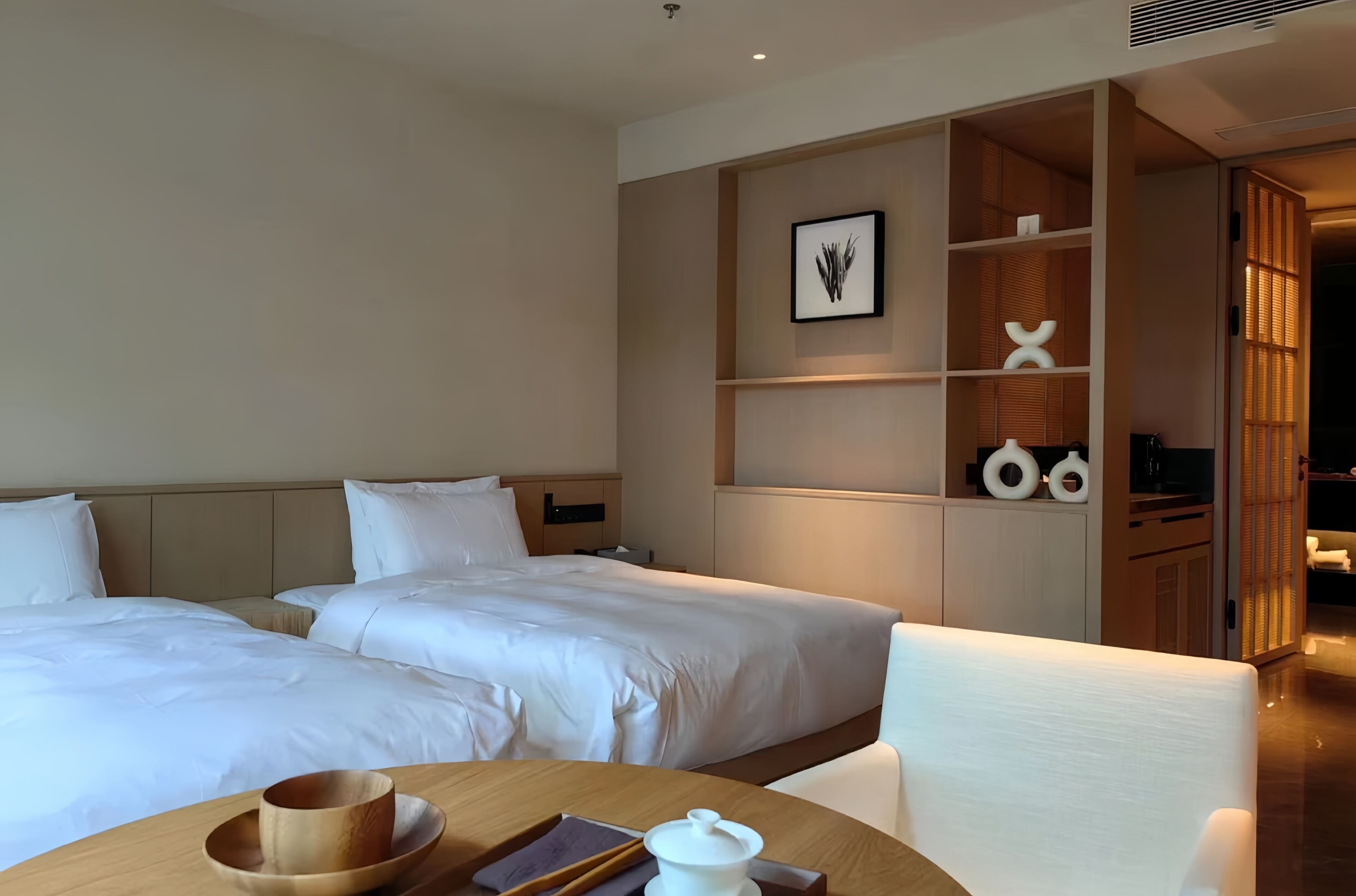 A hotel room with two beds, a wooden table with a tray, a white pitcher, and a framed picture on the wall.