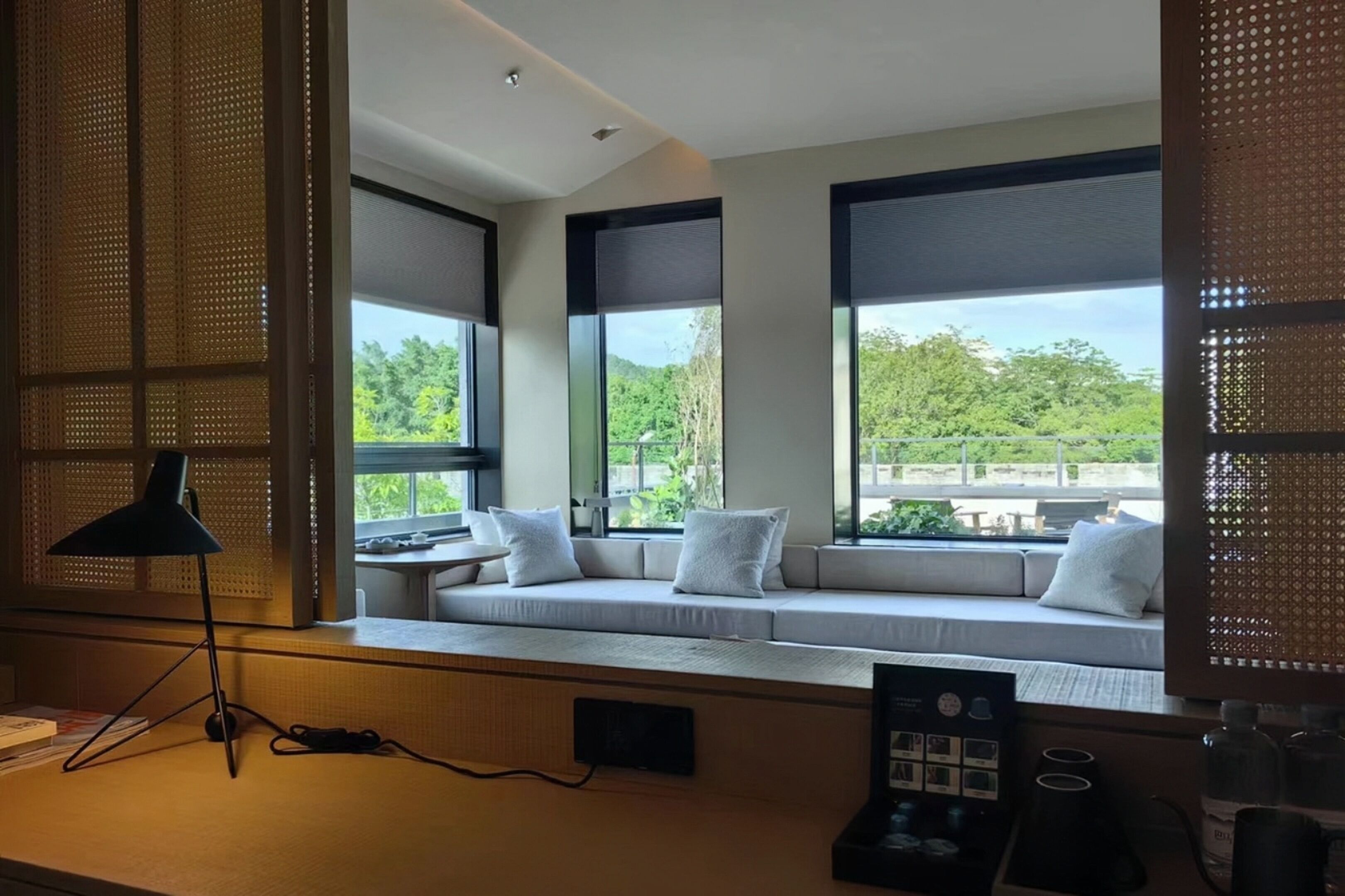 A modern room with a large window seat, a black desk lamp, and a view of greenery outside.