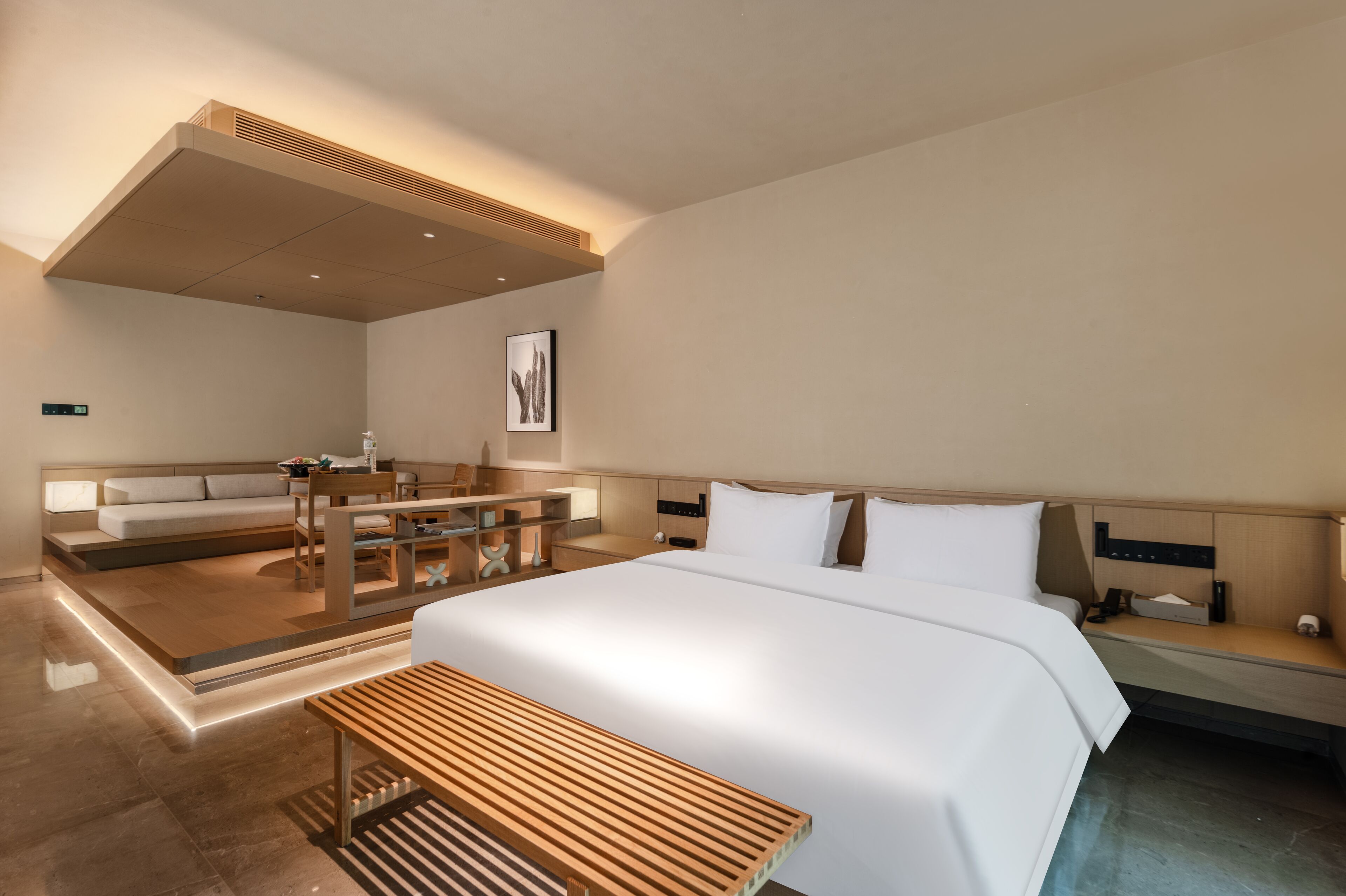 A modern hotel room with a large bed, a wooden bench, a desk, and a seating area with a sofa and chairs.