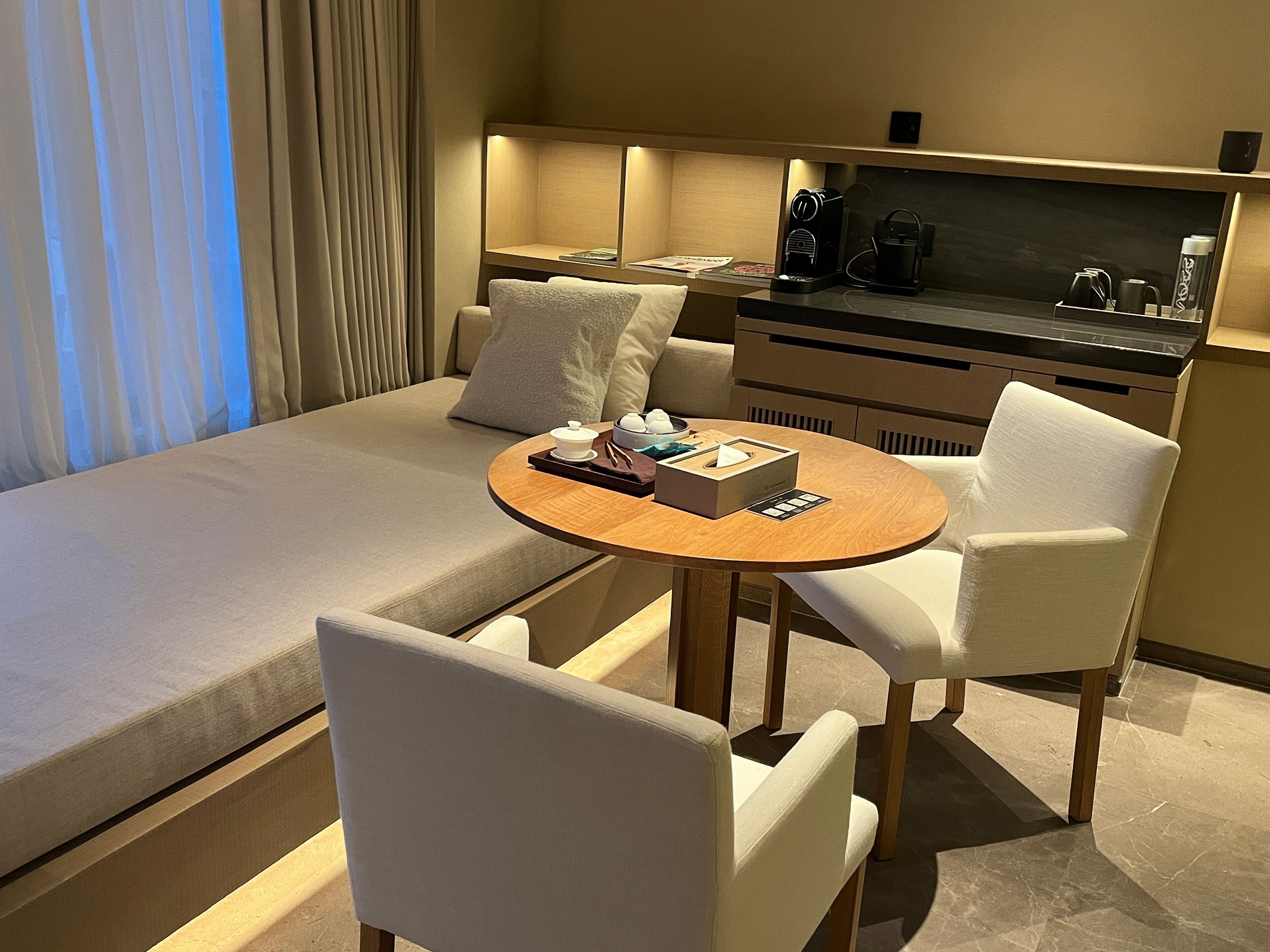 A modern hotel room with a large bed, a round wooden table, two white chairs, a small coffee machine, and a flat-screen TV on a wooden cabinet.