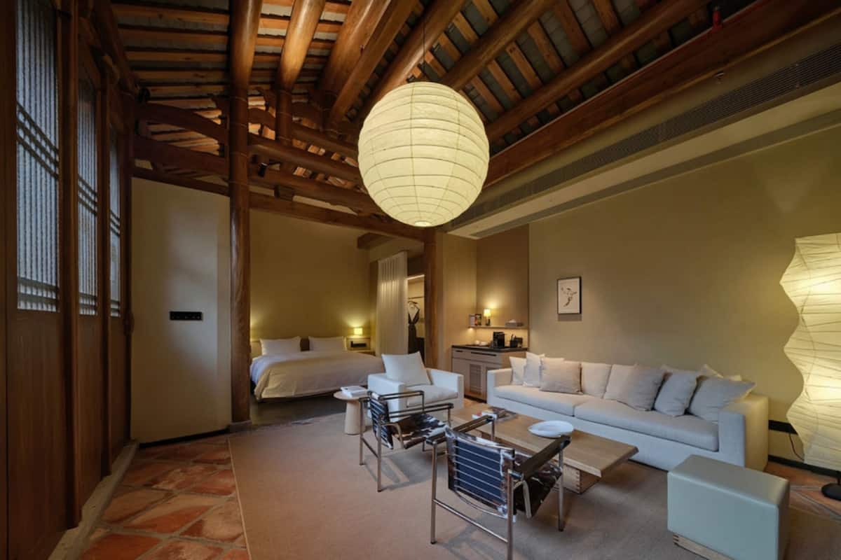 Une chambre d’hôtel avec un plafond en bois, un grand luminaire suspendu, un lit, un canapé, une table basse et une petite table avec des chaises.