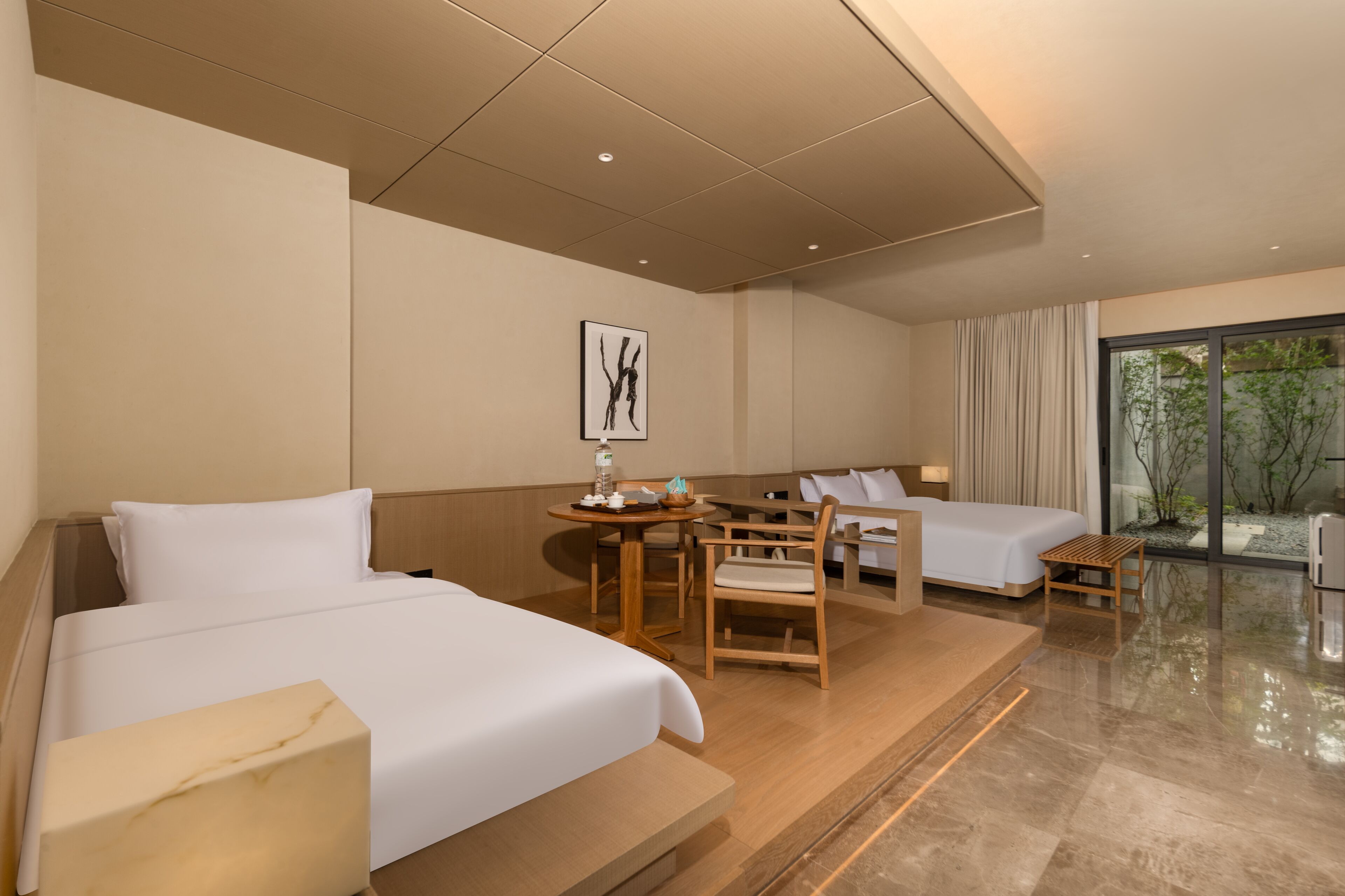 A hotel room with two beds, a dining table, and a view of greenery outside.