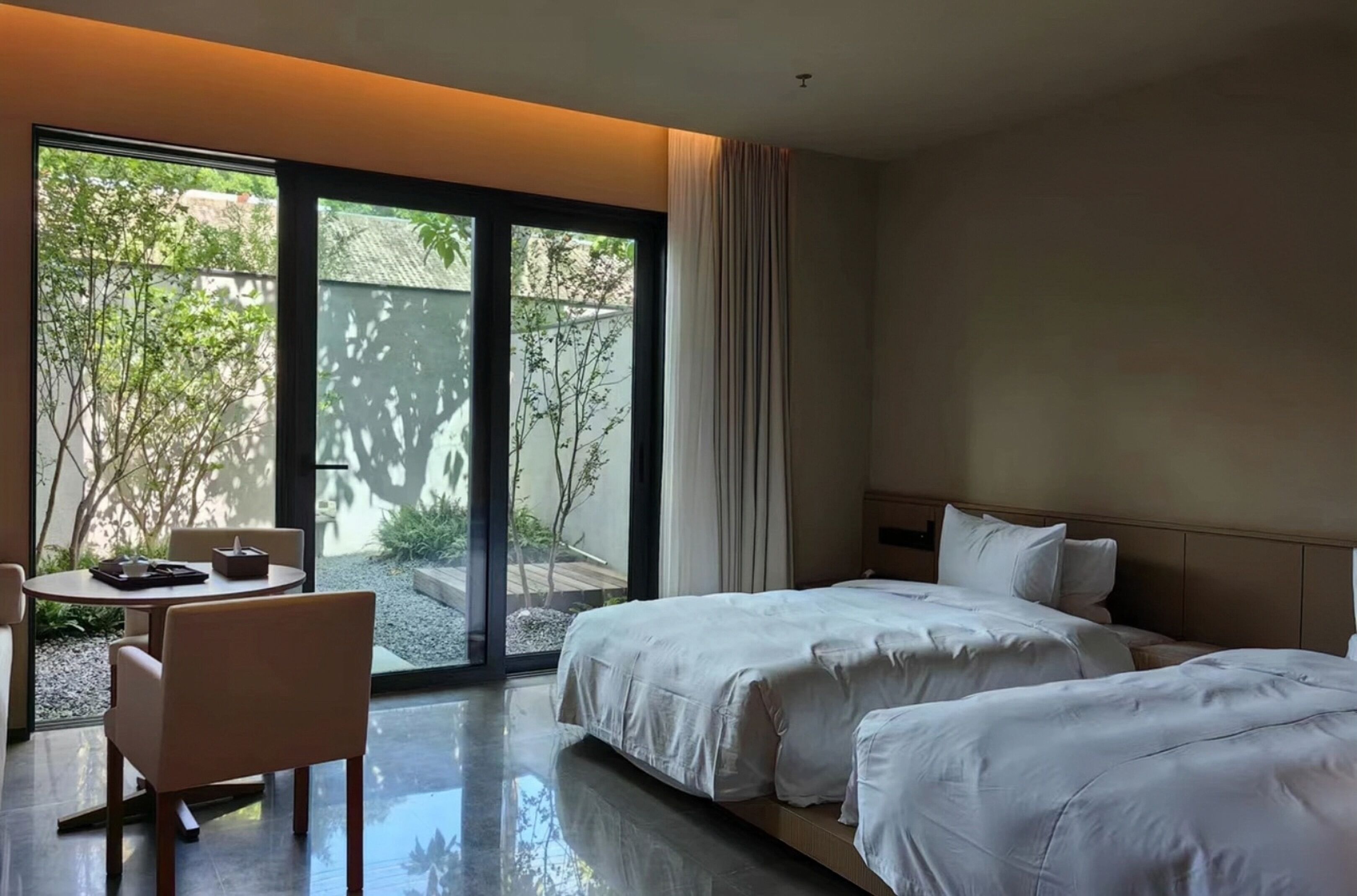 Una camera d'albergo con due letti, un tavolino con sedie e un'ampia porta-finestra scorrevole che dà su uno spazio esterno con piante.
