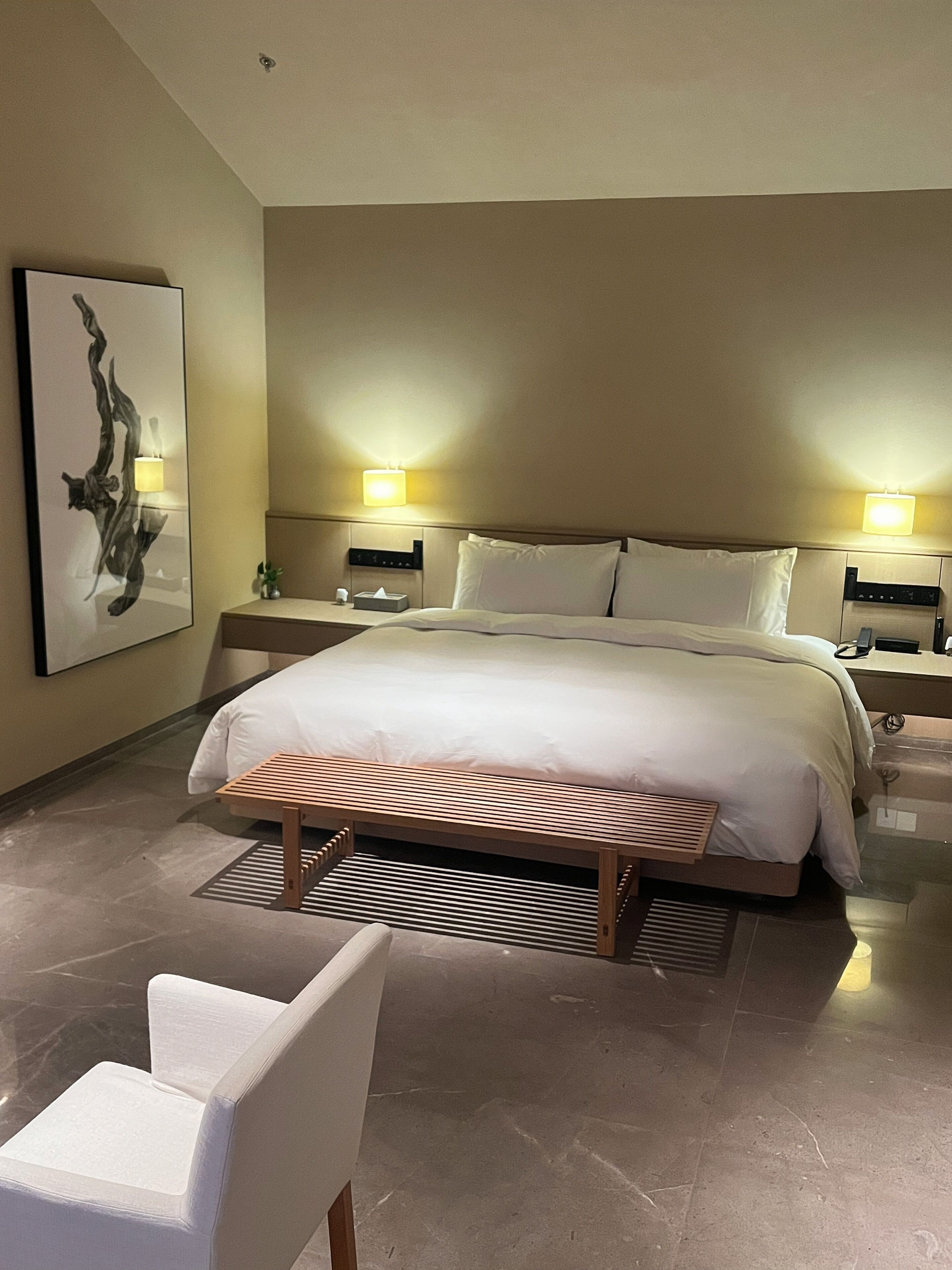 A modern hotel room with a large bed, two bedside lamps, a bedside table, a wall-mounted artwork, and a bench.