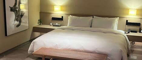 A modern hotel room with a large bed, two bedside lamps, a bedside table, a wall-mounted artwork, and a bench.