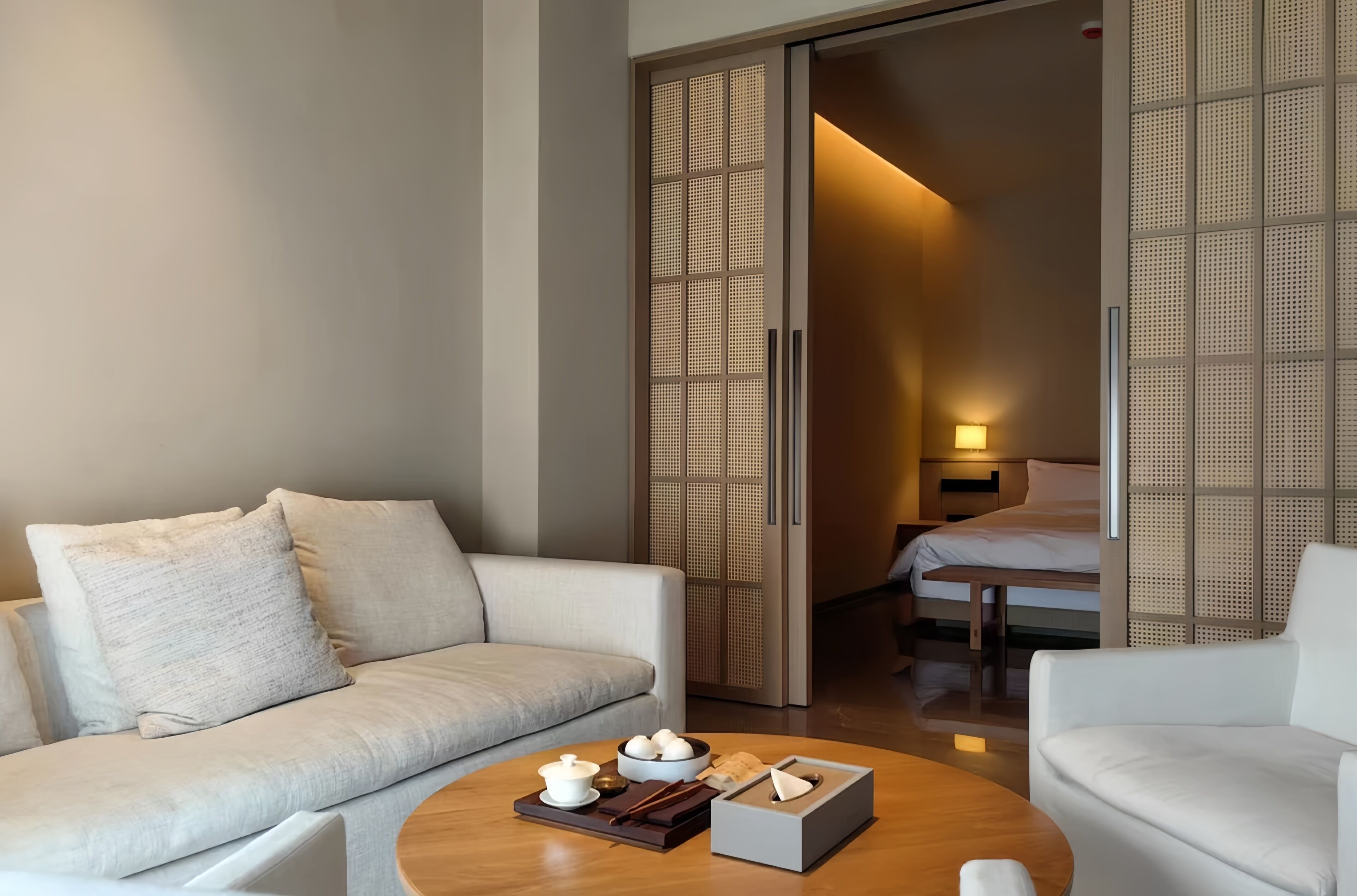 A modern hotel room with a sofa, a round coffee table with tea set, and a bed with a wooden headboard.