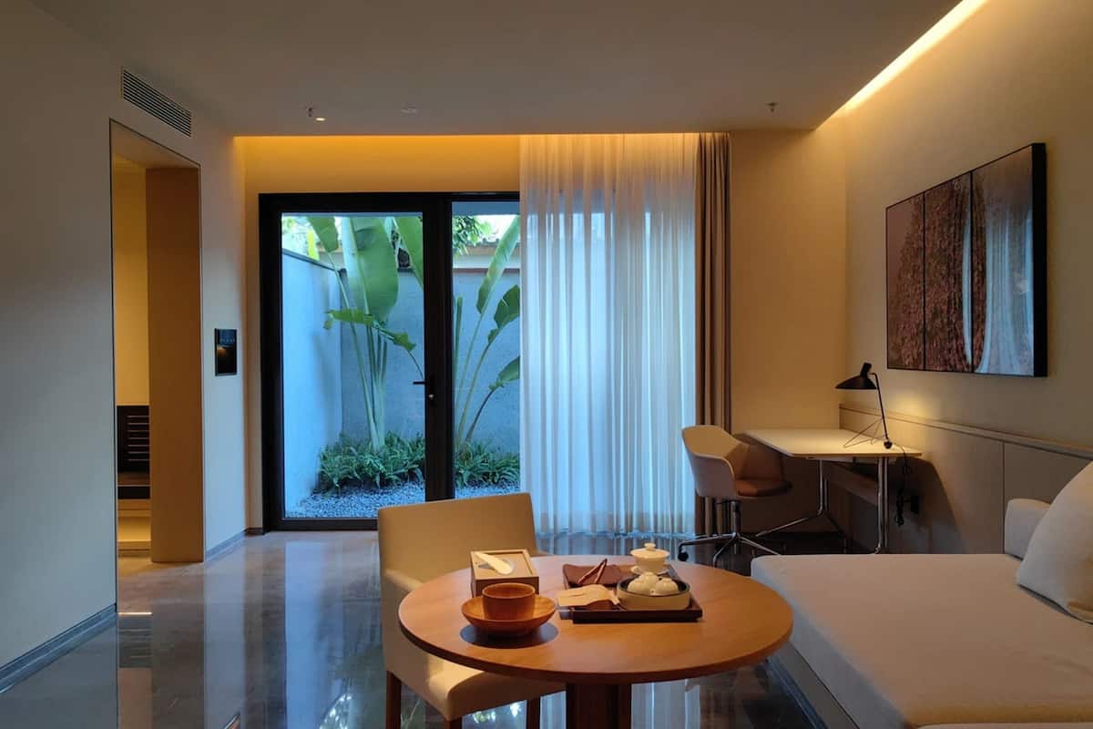Une chambre d’hôtel moderne avec une table à manger ronde, un lit, un bureau et une grande fenêtre avec des rideaux.