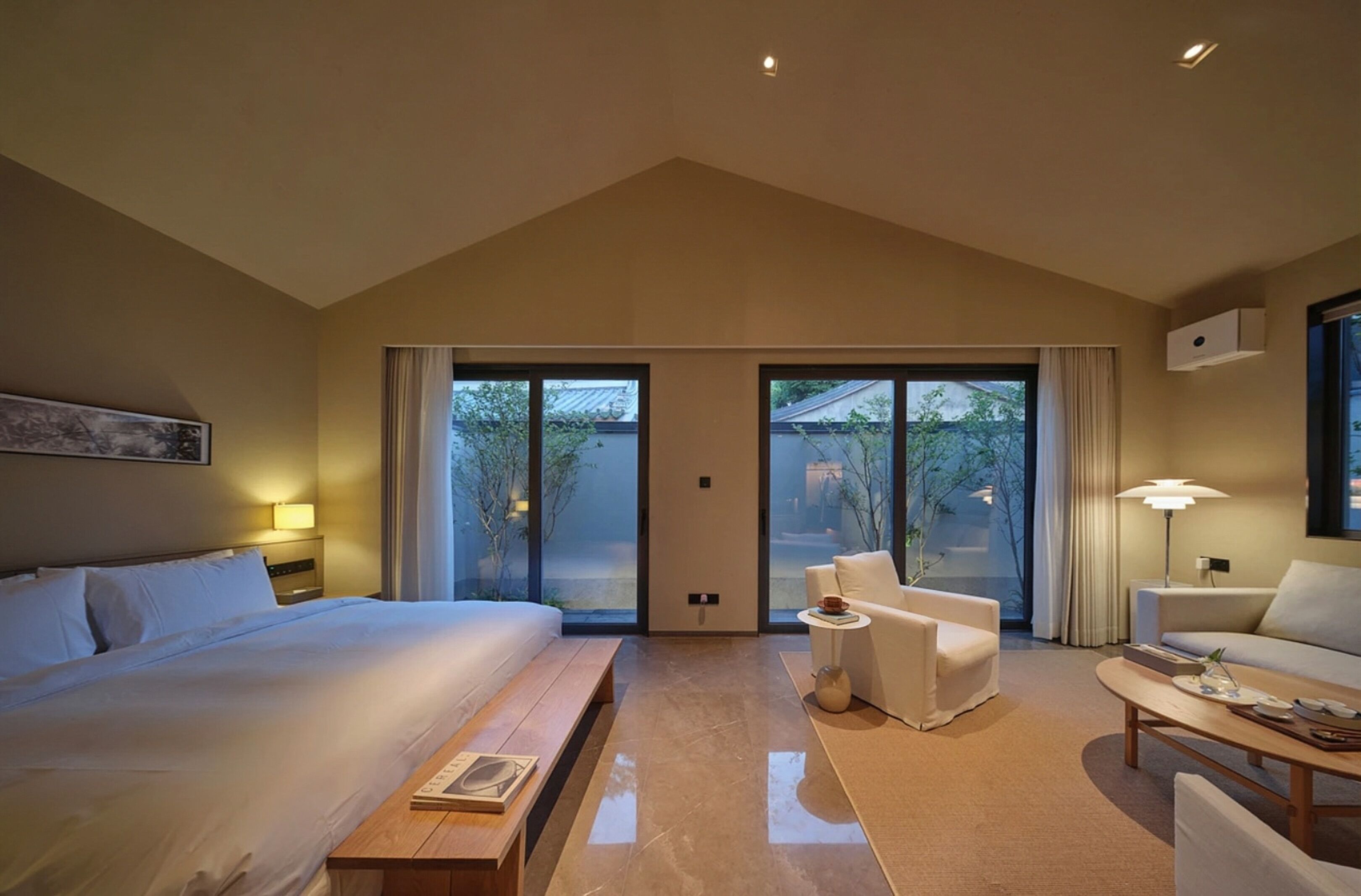 Een moderne hotelkamer met een groot bed, een bank, een salontafel en een bureau. Er zijn grote ramen met gordijnen, uitzicht op het omliggende landschap en een plafondlamp.