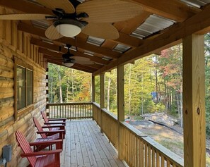 En stuga av trä med en veranda, röda Adirondack-stolar och utsikt över en skog.