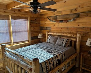 Un dormitorio de cabaña de madera con una cama, un ventilador de techo, una ventana con mosquitero y una pequeña mesa con lámpara.