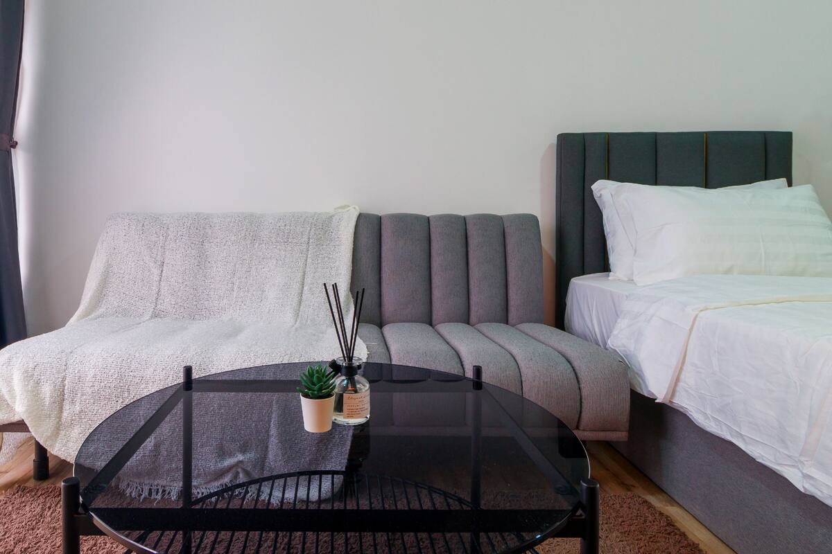 Ein modernes Zimmer mit einem Glastisch, einem grauen Sofa und einem Bett mit weißer Bettwäsche.