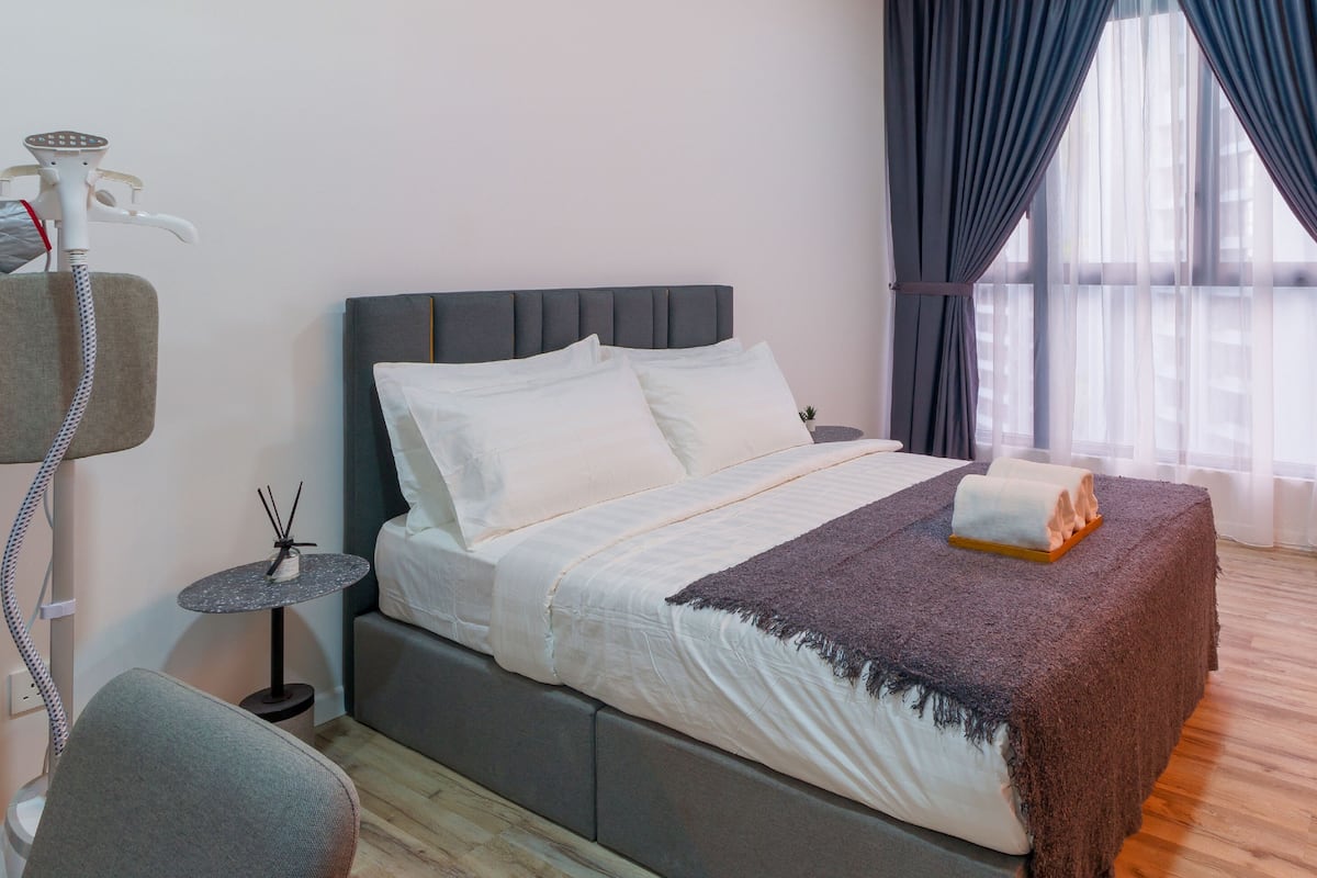 Ein modernes Hotelzimmer mit Bett, einem kleinen Tisch mit Lampe und einem Fenster mit Vorhängen.