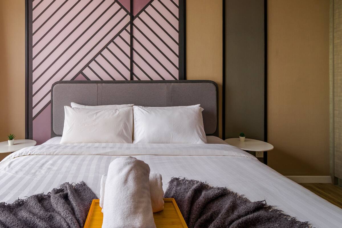 Ein ordentlich bezogenes Bett mit weißen Leinen und Kissen, ein grauer Kopfteil und eine Wand mit geometrischem Muster im Hintergrund.