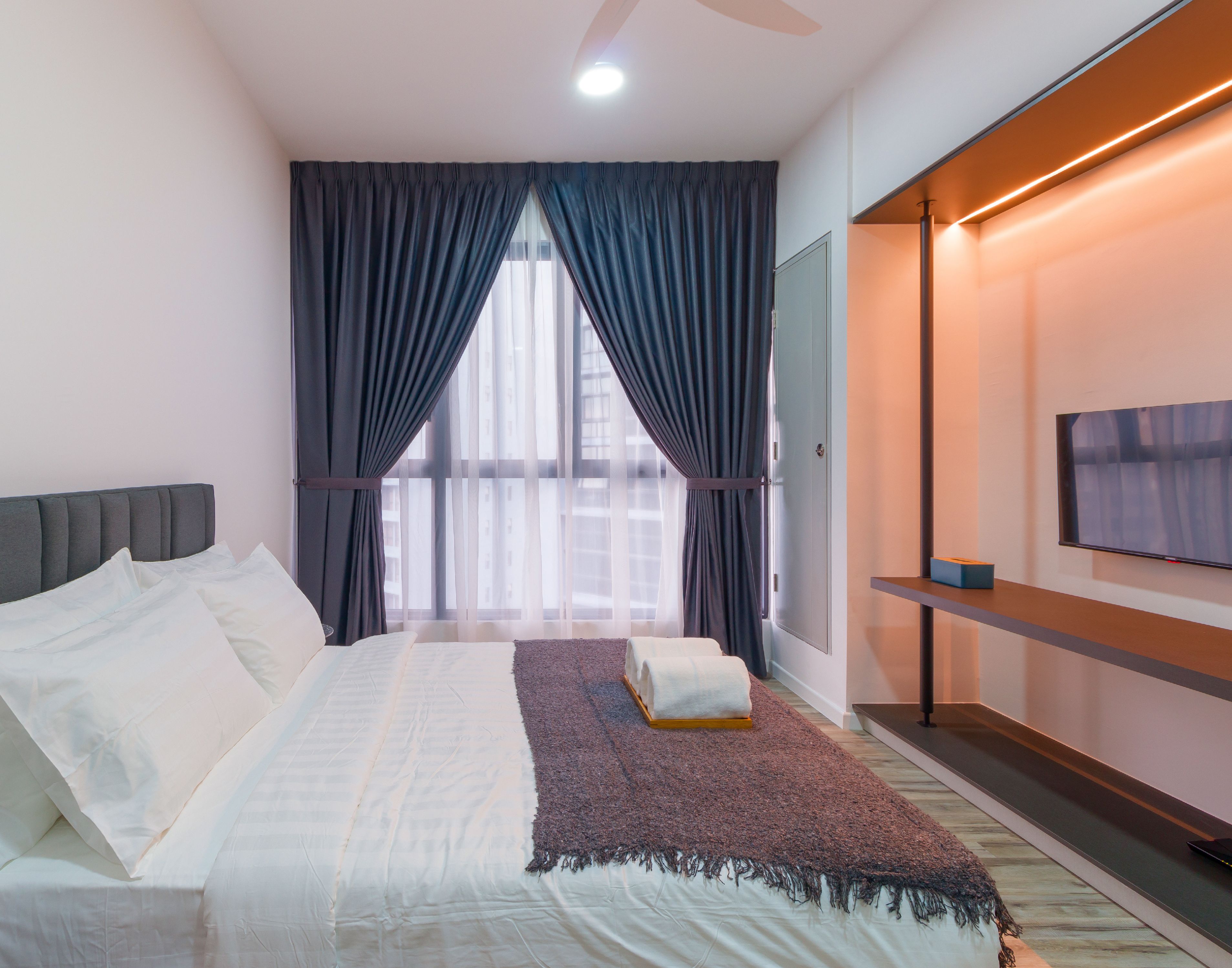 Una moderna camera d'albergo con un letto grande, una TV a schermo piatto, un ventilatore a soffitto e un'ampia finestra con tende.