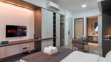 A modern hotel room with a bed, a TV mounted on the wall, a desk with a chair, and a kitchenette.