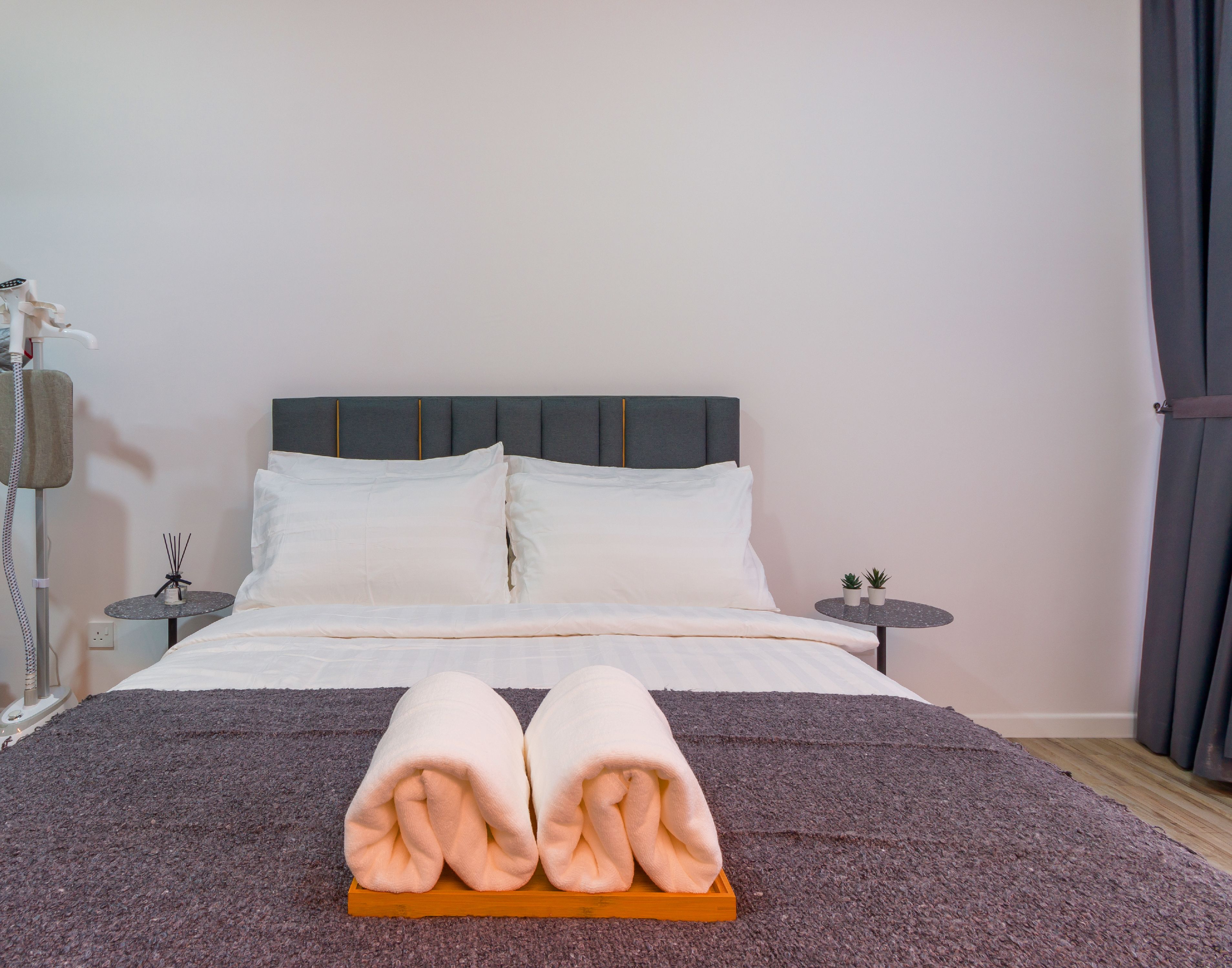 A neatly made bed with a pair of folded towels on a tray, a bedside table with a lamp, and a small table with two potted plants.