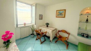 A room with a large window, a table with chairs, a small table with a plant, a picture on the wall, and a shelf with bottles.