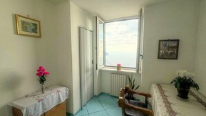 A small room with a window, a radiator, a wooden chair, a bedside table with a vase of flowers, and a bed with a floral bedspread.