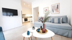 Una sala de estar moderna con un sofá gris, un televisor de pantalla plana montado en la pared y un pequeño comedor con una mesa y sillas blancas.