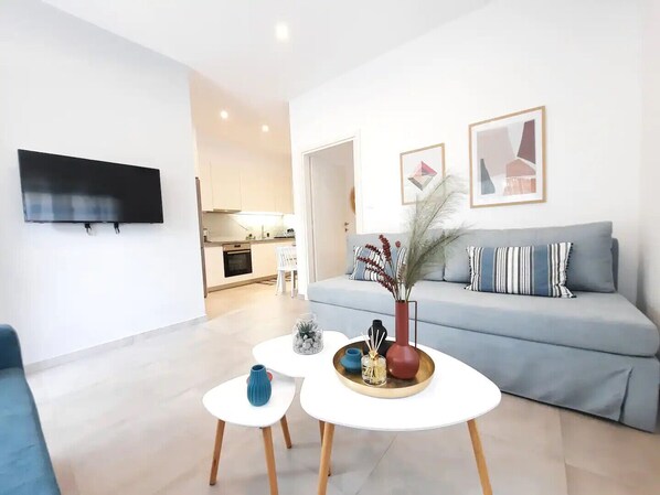 A modern living room with a grey sofa, a flat-screen TV mounted on the wall, and a small dining area with a white table and chairs.