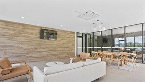 A modern lounge area with a wooden wall, a white sofa, a dining table with chairs, and a large window offering a view. - 2 Bedroom Luxury in Burleigh Heads! (Gold Coast)
