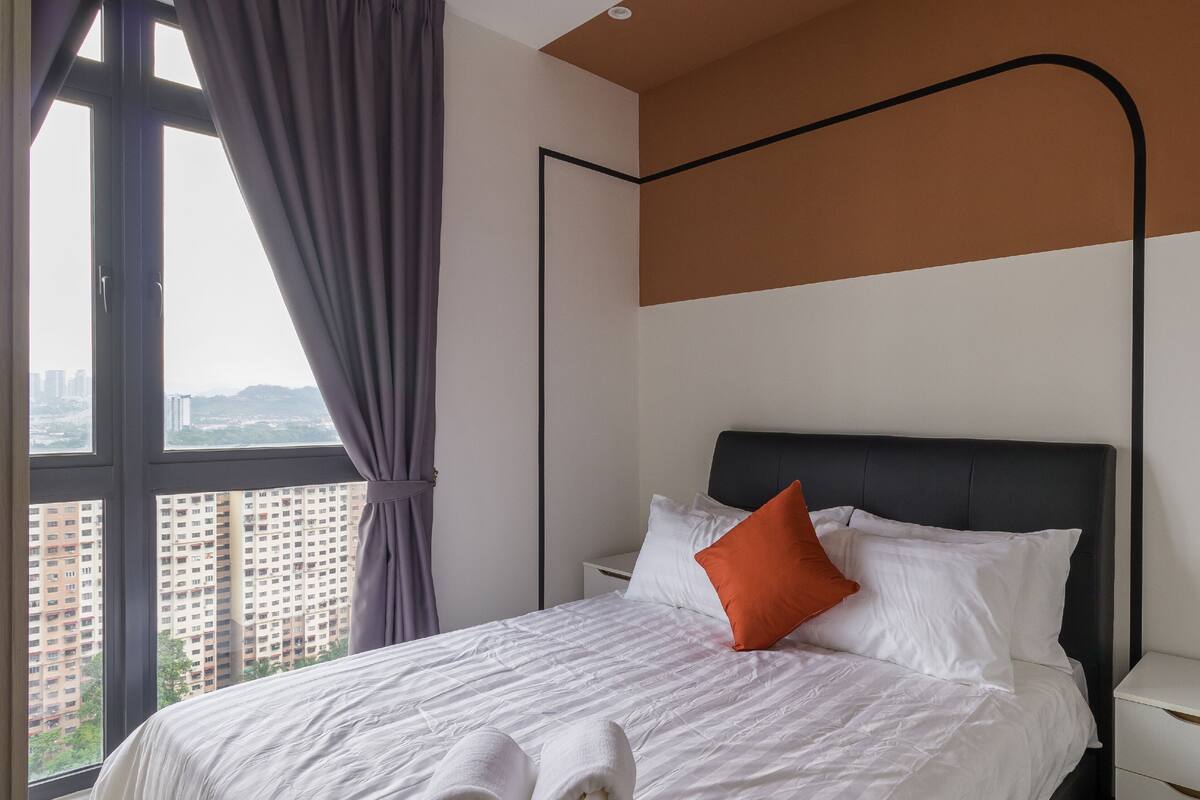 Une chambre d’hôtel moderne avec un grand lit, du linge de lit blanc, une tête de lit noire et une vue sur le paysage urbain, visible à travers de grandes fenêtres.