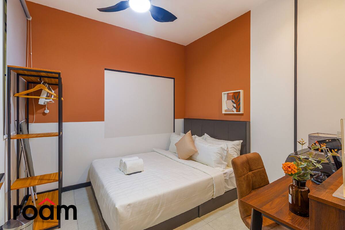 Une chambre d’hôtel équipée d’un lit, d’un bureau, d’une chaise, d’une lampe de bureau, d’un vase avec des fleurs, d’un ventilateur de plafond et d’un tableau blanc fixé au mur.
