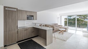 Una cuina moderna amb microones integrat, zona de menjador amb taula i cadires blanques, i un gran finestral corredís que dona a un balcó amb vistes a zones verdes.