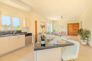 A modern kitchen with a dining area, featuring a large island with a bottle of champagne and glasses, a view of the outdoors, and a ceiling fan.