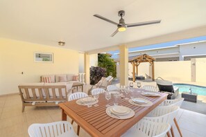Ein Essbereich mit einem Holztisch, gedeckt für sechs Personen, weißen Stühlen, einem Deckenventilator und Blick auf einen Swimmingpool.
