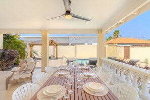 A covered outdoor dining area with a table set for a meal, surrounded by white chairs.
