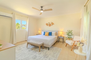 A bedroom with a bed, bedside tables, a bench, a chair, a ceiling fan, and a window.