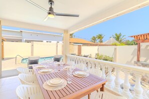 Ein überdachter Essbereich im Freien mit Pool, weißen Stühlen und einem Holztisch, gedeckt für eine Mahlzeit.